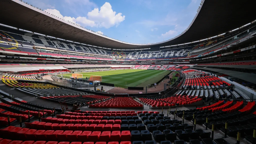 Vista panorámica desde las gradas del Estadio Banorte, mostrando las butacas rojas en primer plano, el terreno de juego verde y el escudo del Club América al fondo bajo un cielo azul.