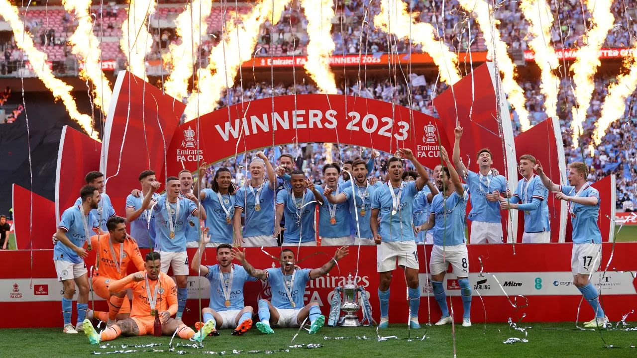 MANCHESTER CITY Jugadores del Manchester City celebrando el título de la FA Cup 2023 en el estadio de Wembley, con fuegos artificiales y el trofeo en el centro bajo un arco de "WINNERS 2023".