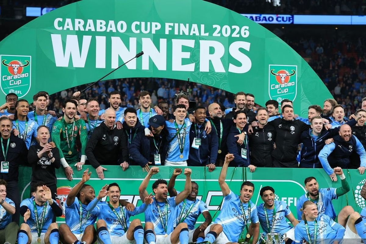 MANCHESTER CITY Jugadores y cuerpo técnico del Manchester City celebrando el título de la Carabao Cup 2026 bajo un arco verde de "WINNERS", con Phil Foden y Pep Guardiola en el centro.