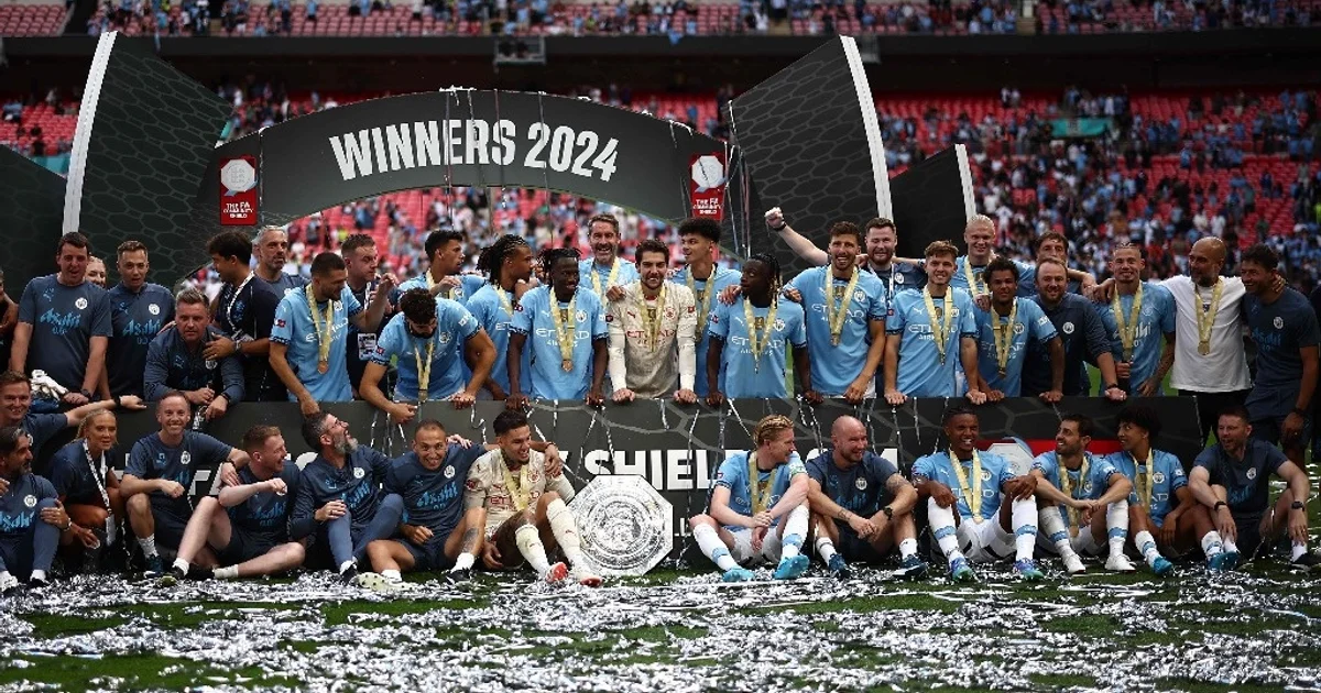 MANCHESTER CITY Jugadores del Manchester City celebrando el título de la Community Shield 2024 en el estadio de Wembley, con el trofeo plateado en el centro y confeti bajo un arco de "WINNERS 2024".