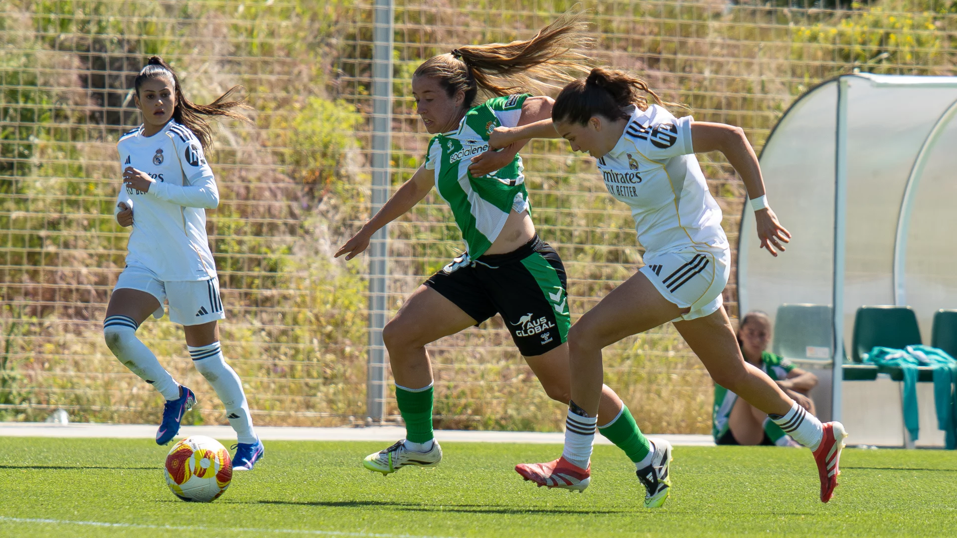 BETIS Fotografía de acción de un partido de fútbol femenino. Una jugadora del Real Betis Féminas con uniforme verde y blanco corre controlando el balón mientras otra jugadora del Real Madrid, con uniforme blanco, la marca muy de cerca. Al fondo, a la izquierda, otra jugadora del Real Madrid observa la jugada.