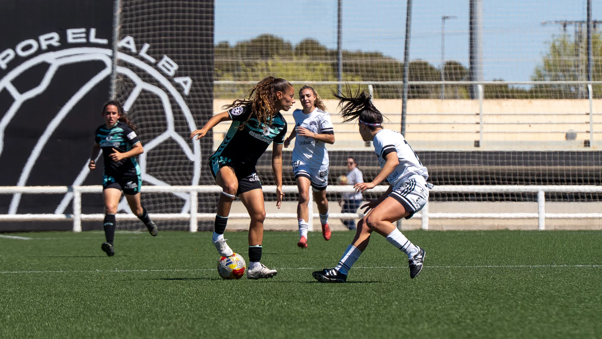 BETIS Fotografía de acción de un partido de fútbol femenino. Una jugadora del Real Betis Féminas, con uniforme negro y detalles turquesas, pisa el balón con su pie derecho mientras es marcada por una jugadora del equipo rival vestida de blanco. Al fondo se ven otras dos jugadoras y una valla publicitaria grande que dice "POR EL ALBA".