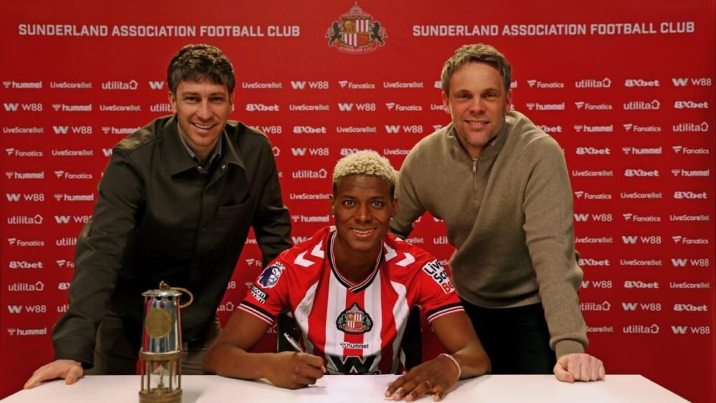 Jugador juvenil Nilson Angulo firmando como uno de los fichajes de el Sunderland AFC acompañado de directivos frente al panel de patrocinadores.
