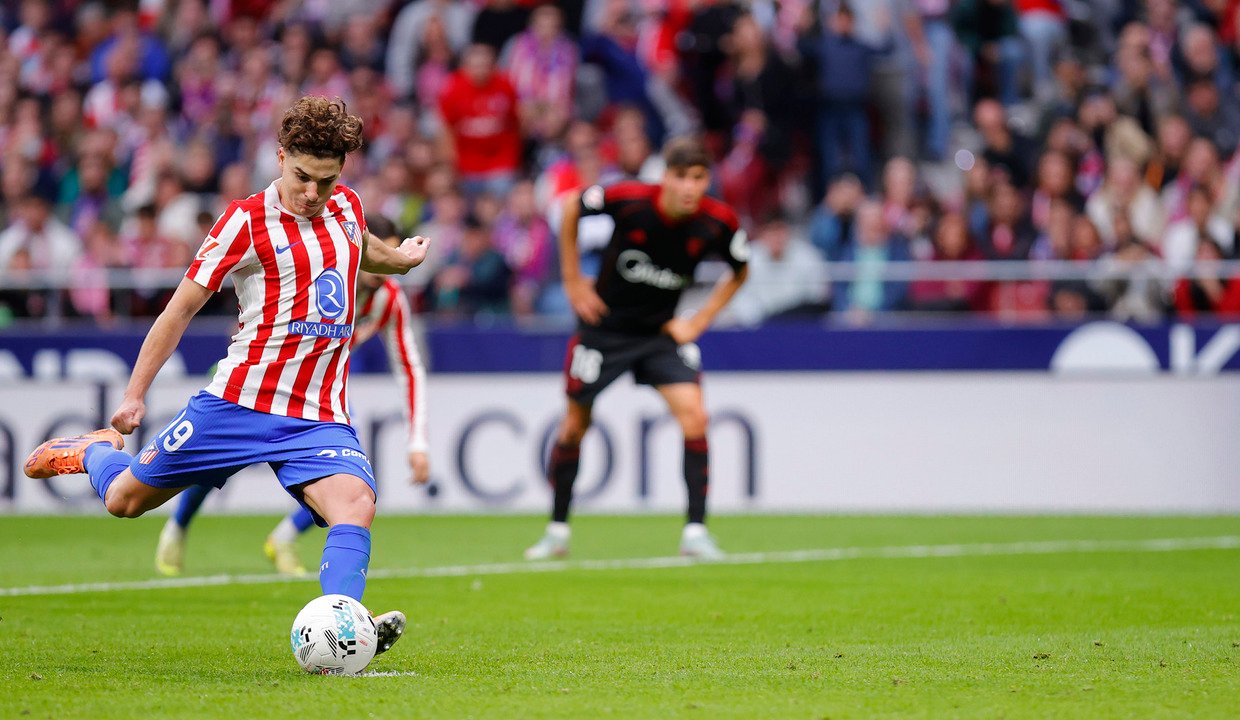 Fotografía de acción de Julián Álvarez pateando un balón de fútbol con la pierna derecha. Viste la equipación local del Atlético de Madrid con pantalones azules y medias azules. De fondo, la grada del estadio llena de aficionados y un jugador rival observando la jugada.