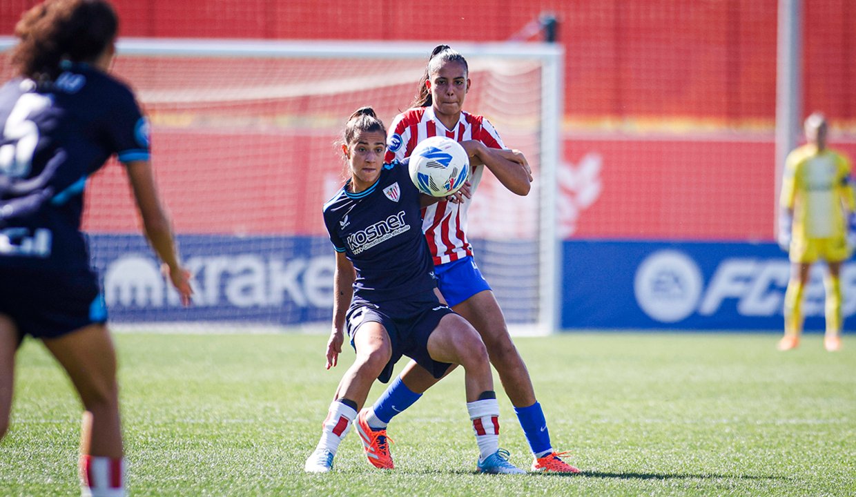Fotografía de acción de Leicy Santos protegiendo el balón con el cuerpo frente a una defensora rival. Viste la camiseta rojiblanca del Atlético de Madrid; la oponente viste un uniforme oscuro con el patrocinador Kosner. De fondo se observa una portería y vallas publicitarias en un día soleado.
