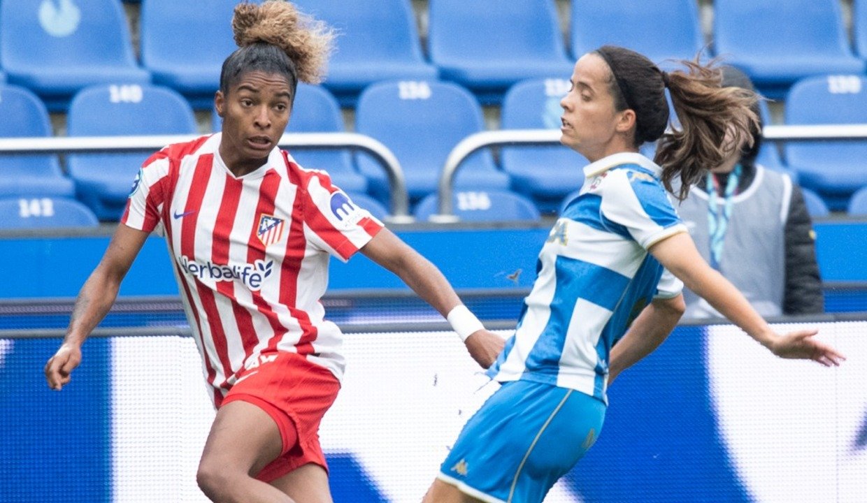 Fotografía de acción de la futbolista Deyna Castellanos con la camiseta rojiblanca del Atlético de Madrid (patrocinio de Herbalife) en un duelo contra una defensora del Deportivo Abanca vestida de azul y blanco. De fondo se aprecian las gradas azules del estadio.