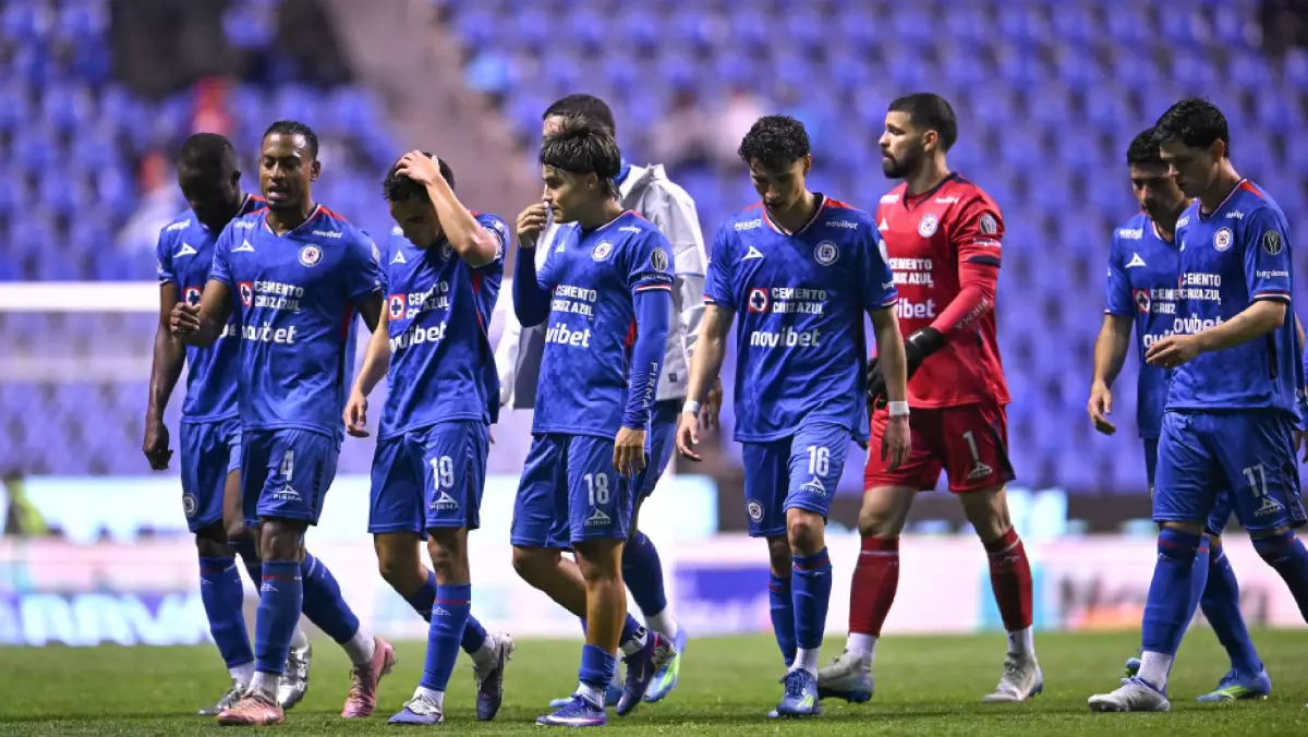 INICIO Jugadores de Cruz Azul caminando por el campo de juego con semblante serio. Se observa a los futbolistas con sus uniformes azules y al portero de rojo, en un ambiente nocturno de estadio.